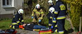 Ćwiczenia w DPS. Na trawniku, na noszach typu deska leży poszkodowany mężczyzna. Bezpośrednio przy nim znajduje się pięciu strażaków udzielających mu kwalifikowanej pierwszej pomocy. Dwaj klęczą - jeden z nich stabilizuje głowę. Obok znajduje się sprzęt ratownictwa medycznego. W tle budynek oraz zieleń parkowa.
