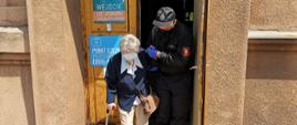 Strażak podtrzymując starszą kobietę pod rękę wychodzi z punktu szczepień. Kobieta podpiera się na kuli.