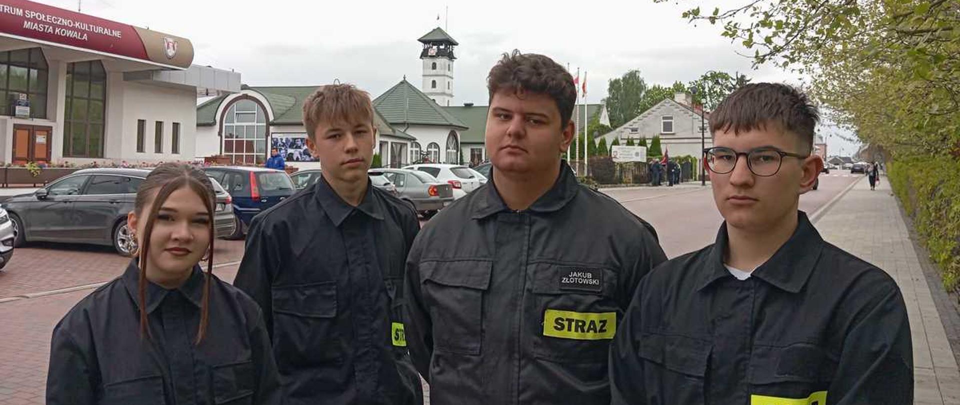 Na tym zdjęciu widzimy czworo młodych ludzi, prawdopodobnie członków Młodzieżowej Drużyny Pożarniczej lub młodych druhów z OSP Kowal. Stoją oni blisko siebie, patrząc w stronę obiektywu, na tle infrastruktury miejskiej.Oto szczegółowy opis:Pierwszy plan (osoby): Od lewej strony stoi dziewczyna o ciemnych włosach spiętych w kucyk, a obok niej trzech chłopców. Wszyscy mają na sobie czarne mundury koszarowe z charakterystycznymi żółtymi emblematami „STRAŻ” na piersi. Chłopak stojący w centrum ma nad napisem przypiętą imienną naszywkę: „JAKUB ZŁOTOWSKI”. Ich twarze wyrażają powagę i skupienie.Tło i architektura:Po lewej stronie, w głębi, widoczny jest nowoczesny budynek z czerwonym dachem i napisem na elewacji: „CENTRUM SPOŁECZNO-KULTURALNE MIASTA KOWALA”.Dokładnie pomiędzy głowami chłopców, w oddali, góruje charakterystyczna biała wieża z galeryjką na szczycie i ciemnym daszkiem – jest to prawdopodobnie wieża strażacka przy tutejszej remizie.Po prawej stronie ciągnie się chodnik z kostki brukowej, a nad nim zwieszają się gałęzie drzew z młodymi, jasnozielonymi liśćmi.Kontekst i atmosfera: Zdjęcie zostało wykonane przy rozproszonym, dziennym świetle (pochmurne niebo). Całość sugeruje, że młodzi strażacy biorą udział w tych samych uroczystościach majowych (Święto Konstytucji 3 Maja i Dzień Strażaka), które widzieliśmy na poprzednich zdjęciach z pomnikiem i w kościele.