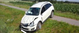 Auto osobowe po zderzeniu z innym pojazdem stoi częściowo w rowie (przód pojazdu) i częściowo na przydrożnej ścieżce rowerowej. Auto ma uszkodzoną przednią maskę, zderzak oraz przednią szybę.