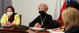 Iwona Michałek siedzi przy stole konferencyjnym. Na twarzy ma maseczkę. Przed sobą trzyma notatki. Obok siedzą osoby z departamentów merytorycznych.