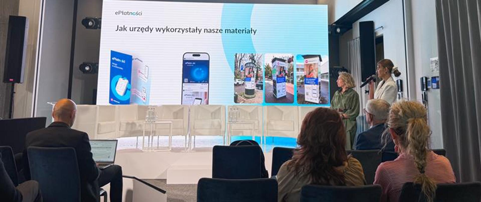 Sala konferencyjna. Na ekranie prezentacja. Na pierwszym planie słuchacze, w tle dwie prezentujące kobiety. 