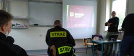 Mężczyzna operaty o parapet prowadzi wykład dla strażaków Państwowej Straży Pożarnej. Przed funkcjonariuszami wyświetlany jest slajd z rzutnika.