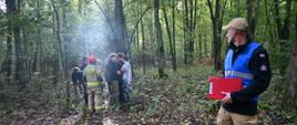 Grupa uczestników ćwiczeń stoi z jednym strażakiem w lesie. Inny strażak w niebieskiej kamizelce z czerwoną teczką w ręce stoi i patrzy w ich kierunku. Za nimi wysokei drzewa. 