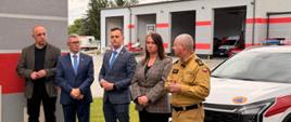 Zdjęcie przedstawia pięć osób stojących przed remizą strażacką. W tle widoczny jest budynek z czerwonymi pasami, otwartymi bramami garażowymi i zaparkowanymi w środku wozami strażackimi. Po prawej stronie znajduje się biały pojazd straży pożarnej. Osoby pozują przed budynkiem; jedna z nich ma na sobie mundur strażacki, a pozostałe są ubrane w stroje cywilne. Nad budynkiem widać pochmurne niebo i drzewa.