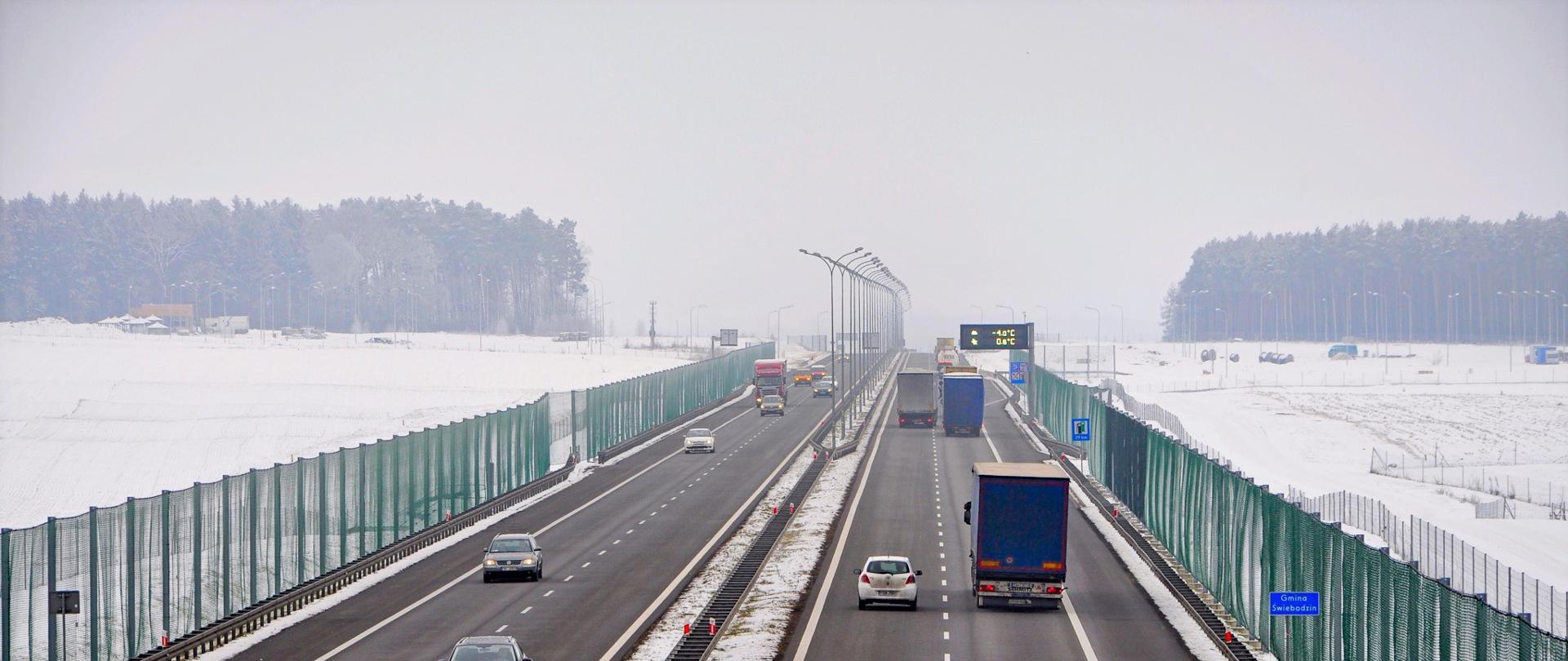 Dwujezdniowa droga szybkiego ruchu. Duży ruch pojazdów osobowych i ciężarowych. Po obu stronach drogi rozwieszone zielone siatki. Na pasie rozdzielającym jezdnie i poza drogą leży śnieg.