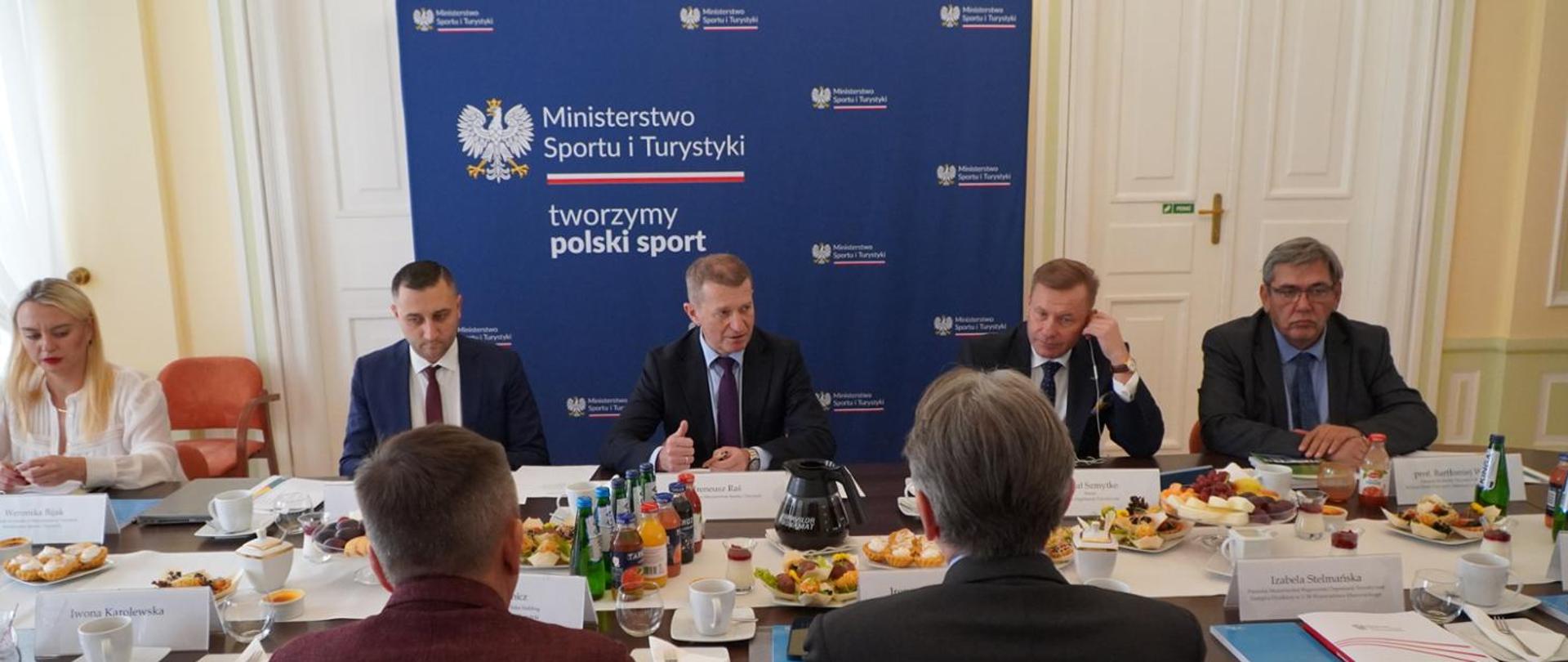 Picture depicting the first meeting of the Tourism Council. People sitting at the table with Minister Raś in the middle. Coffee, tea and snacks on the table. Wall with the Ministry of Sport and Tourism's logos in the background.