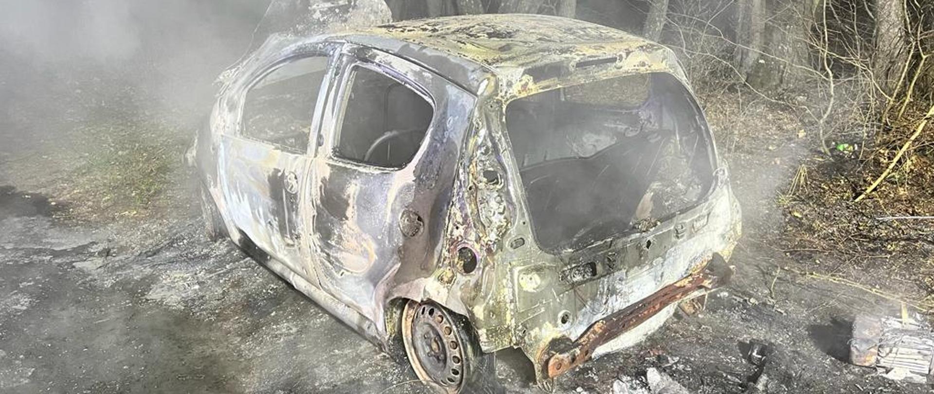 Zdjęcie przedstawia wrak doszczętnie spalonego samochodu osobowego stojący na drodze polnej.