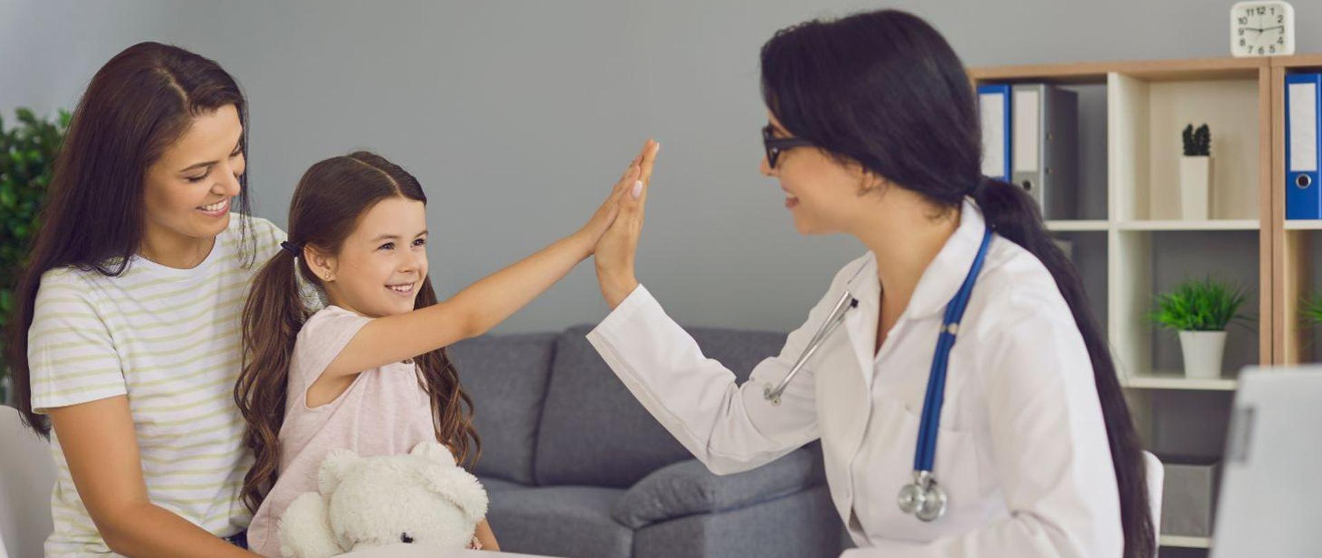 Na zdjęciu z lewej strony uśmiechnięta dziewczynka o długich brązowych włosach z białym misiem siedząca na kolanach mamy przybijająca piątkę z młodą lekarką w okularach. W tle widać siwą sofę, regał z segregatorami i teczkami. 