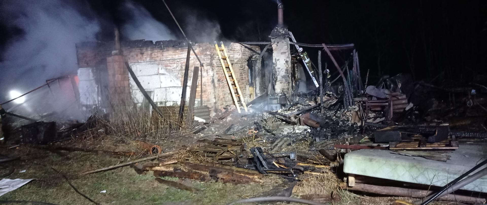 Zdjęcie przedstawia spalony budynek, tło pora nocna. Budynek murowany z czerwonej cegły, dach zawalony. Po prawej stronie stojący komin wokół którego widać spalone elementy drewniane. Przy budynku stoi strażacka drabina drewniana jasno żółtego koloru. Przed budynkiem na gruncie widać węże strażackie. Lewa strona budynku zadymiona, widoczne płomienie wewnątrz budynku. koloru pomarańczowego, natomiast prawa część szaro – żółtego.