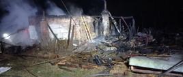 Zdjęcie przedstawia spalony budynek, tło pora nocna. Budynek murowany z czerwonej cegły, dach zawalony. Po prawej stronie stojący komin wokół którego widać spalone elementy drewniane. Przy budynku stoi strażacka drabina drewniana jasno żółtego koloru. Przed budynkiem na gruncie widać węże strażackie. Lewa strona budynku zadymiona, widoczne płomienie wewnątrz budynku. koloru pomarańczowego, natomiast prawa część szaro – żółtego.