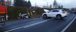 Zdjęcie przedstawia zderzenie dwóch samochodów. Jedno z aut Lexus koloru białego stoi na środku drogi blokując dwa pasy jezdni. Drugie auto znajduje się w przydrożnym rowie. Samochody mają uszkodzone przody. Na zdjęciu widać również strażaków oraz karetkę pogotowia ratunkowego. 