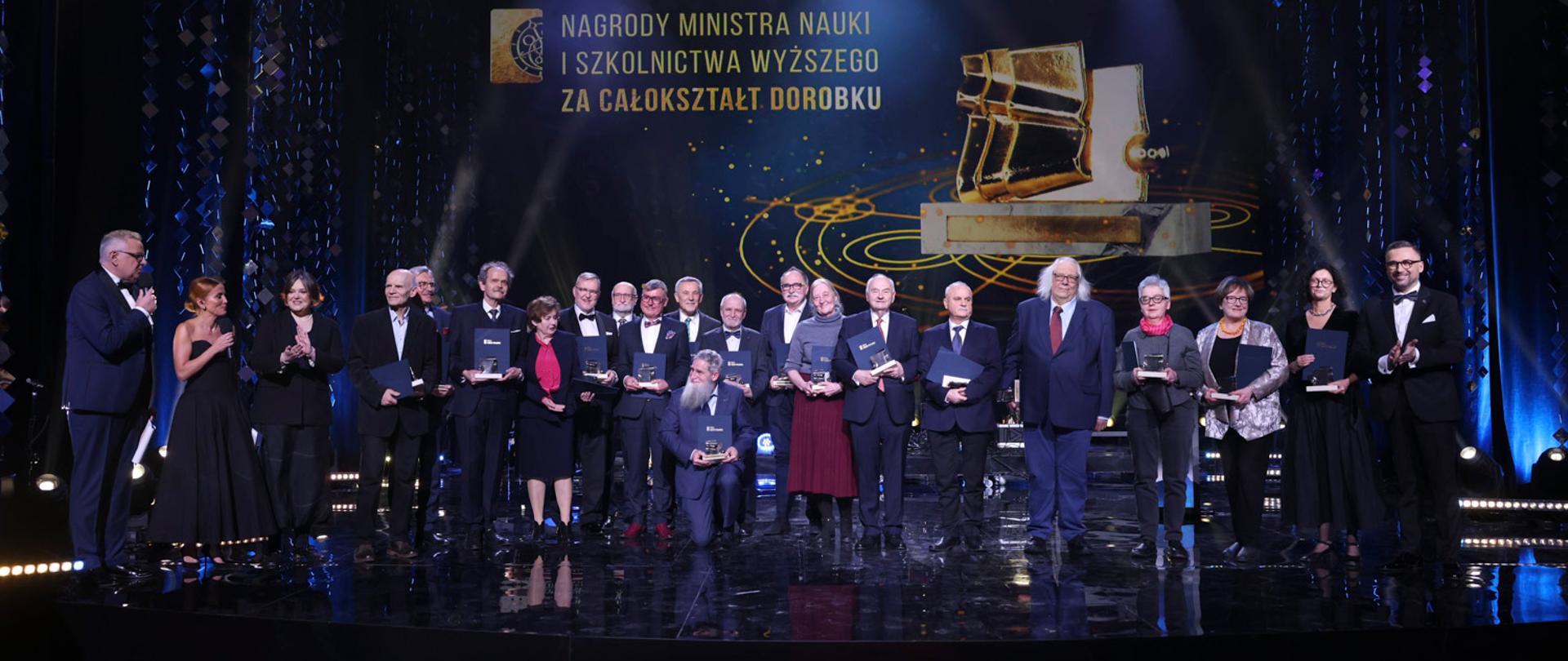 Na podwyższeniu z przodu sali stoi grupa kilkudziesięciu osób trzymających granatowe teczki, za nimi na granatowej ścianie napis Nagrody ministra nauki i szkolnictwa wyższego za całokształt dorobku.