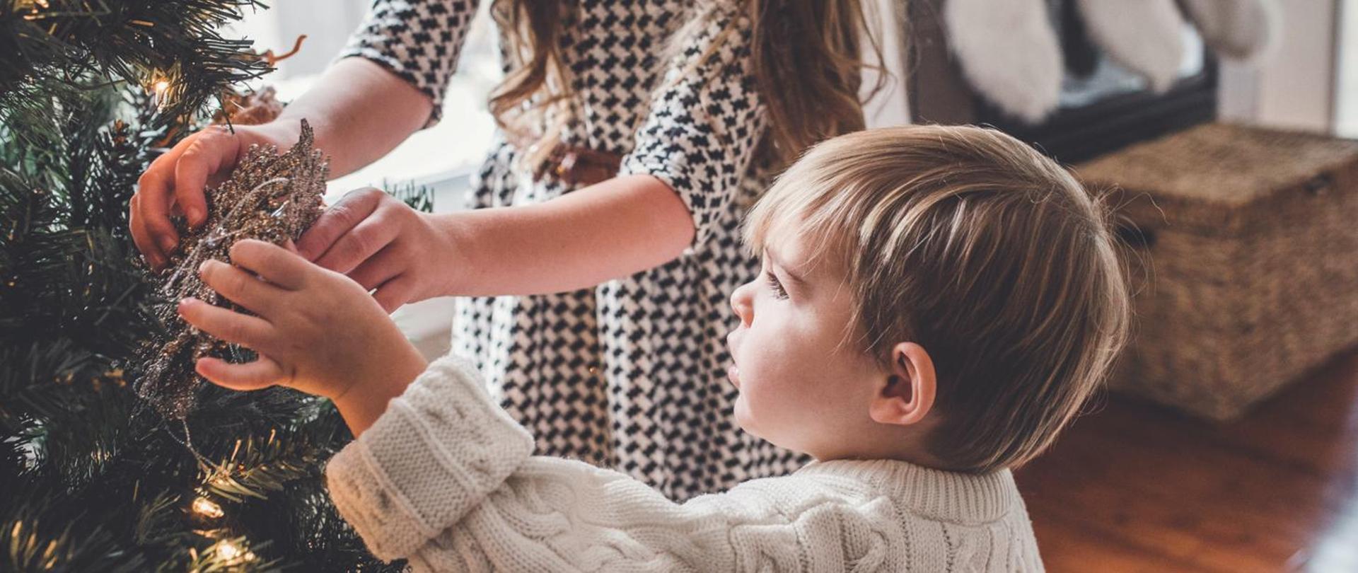 Z lewej strony widać fragment choinki. Na fotografii przedstawiono chłopczyka w białym sweterku z mamą w sukience w kratkę, którzy razem zakładają ozdobę świąteczną na choinkę. W tle wyposażenie pokoju: z lewej strony beżowa skrzynia na brązowej podłodze. 