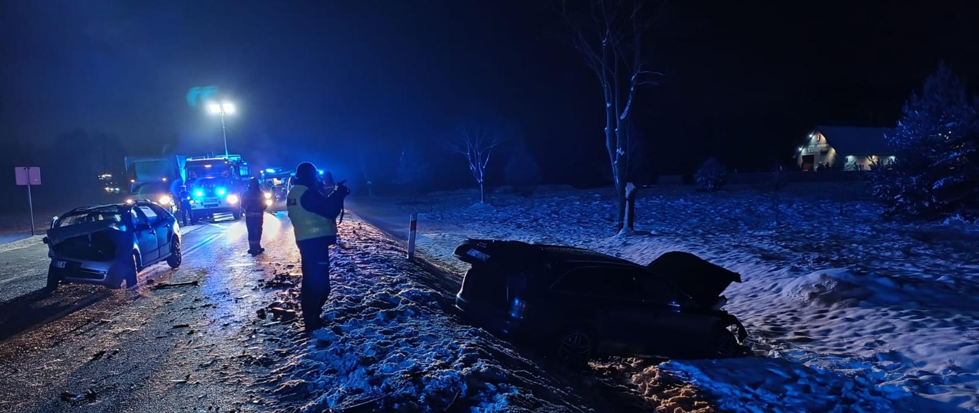 Zdjęcie przedstawia drogę krajowa numer 74. Po prawej stronie w przydrożnym rowie znajduje samochód osobowy, a po lewej, na drodze stoi drugie uszkodzone auto. W głębi widać samochody służbowe oraz wystawiony maszt oświetlający z najaśnicami.