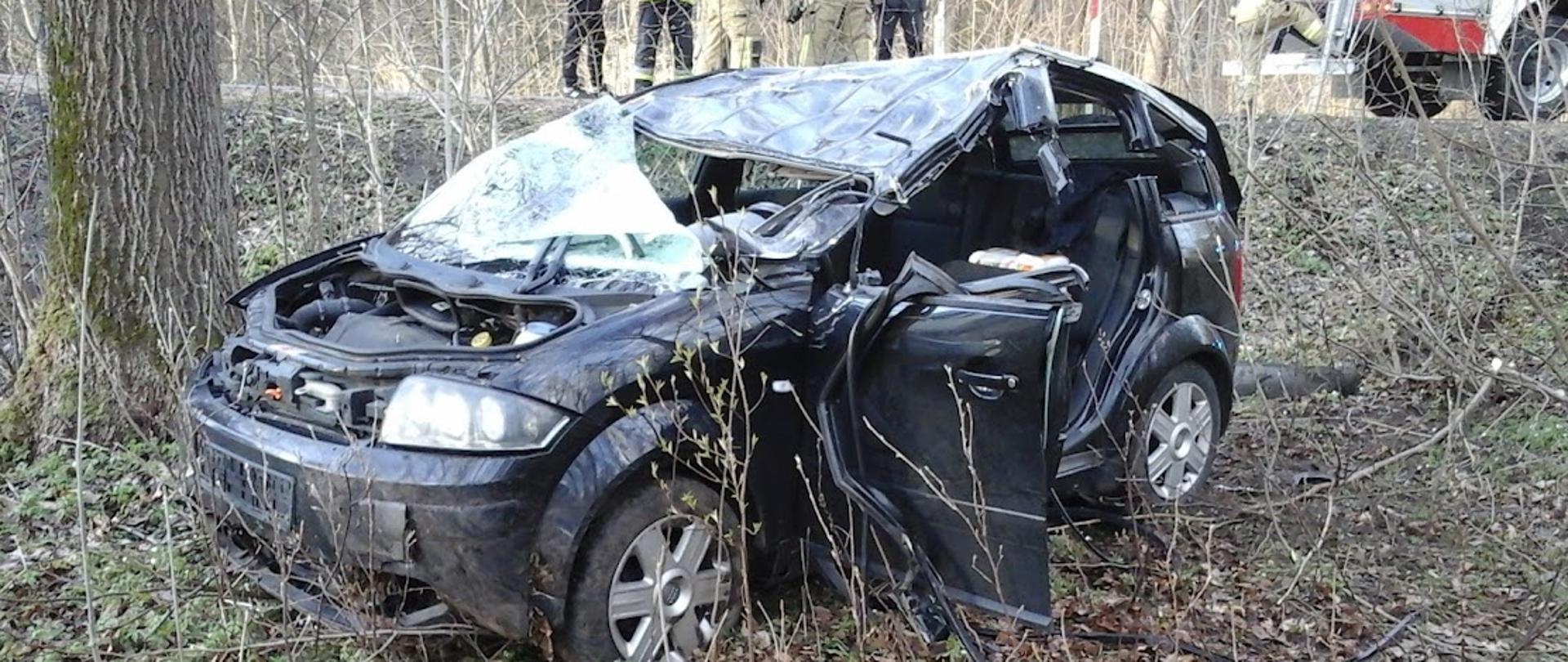 Samochód osobowy po wypadku drogowym stoi w przydrożnym rowie. W aucie zniszczony jest dach, przednia maska i przednia szyba, wycięty został środkowy słupek i drzwi tylne od strony kierowcy.