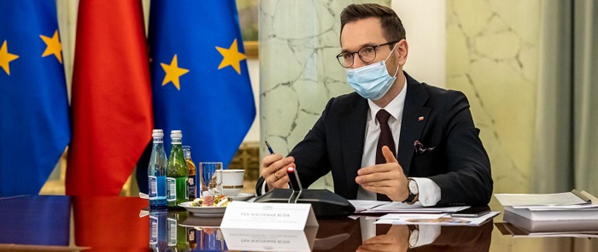 Wiceminister Waldemar Buda siedzi przy stole konferencyjnym, przed nim dokumenty i mikrofon, za nim flagi Polski i Unii Europejskiej