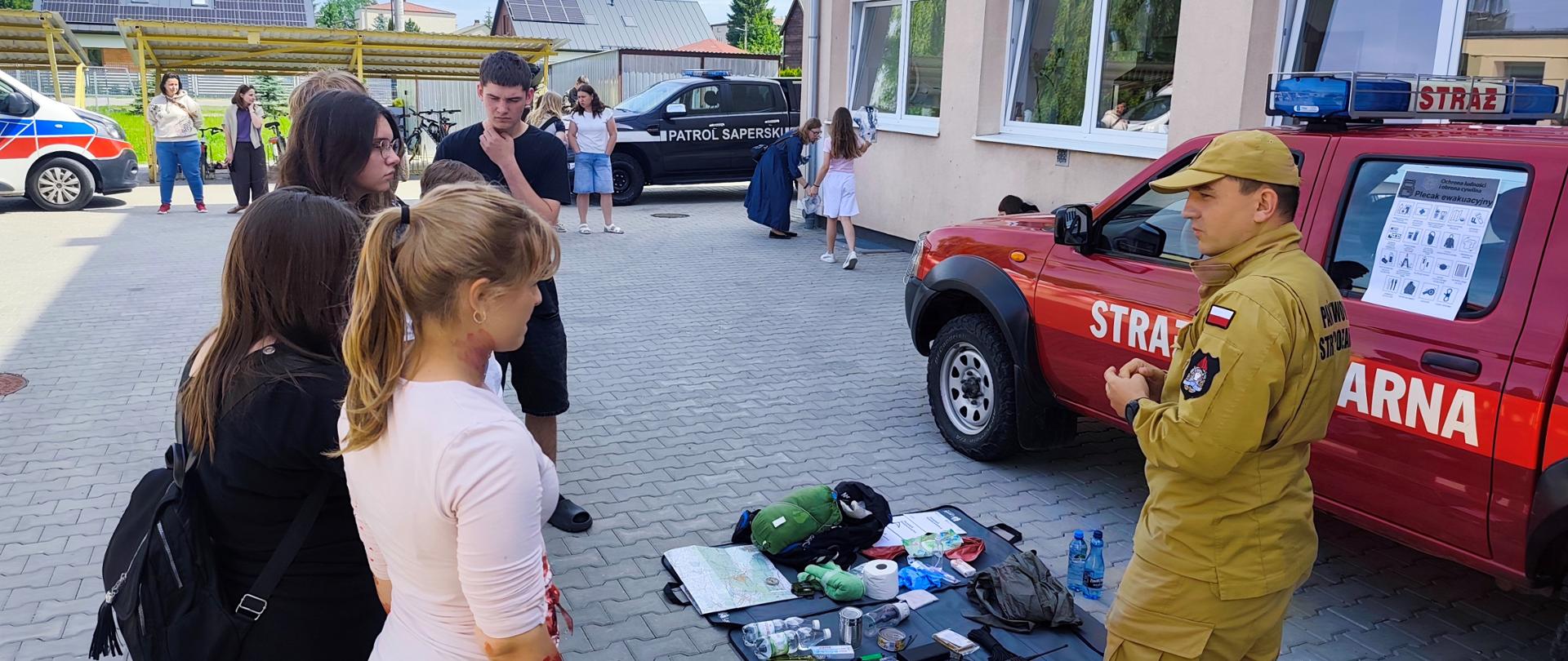 Po prawej stronie fotografii na tle czerwonego strażackiego samochodu operacyjnego typu pick up w żółtym ubraniu służbowym stoi funkcjonariusz PSP, po lewej stronie grupa nastolatków słucha prezentacji strażaka. 