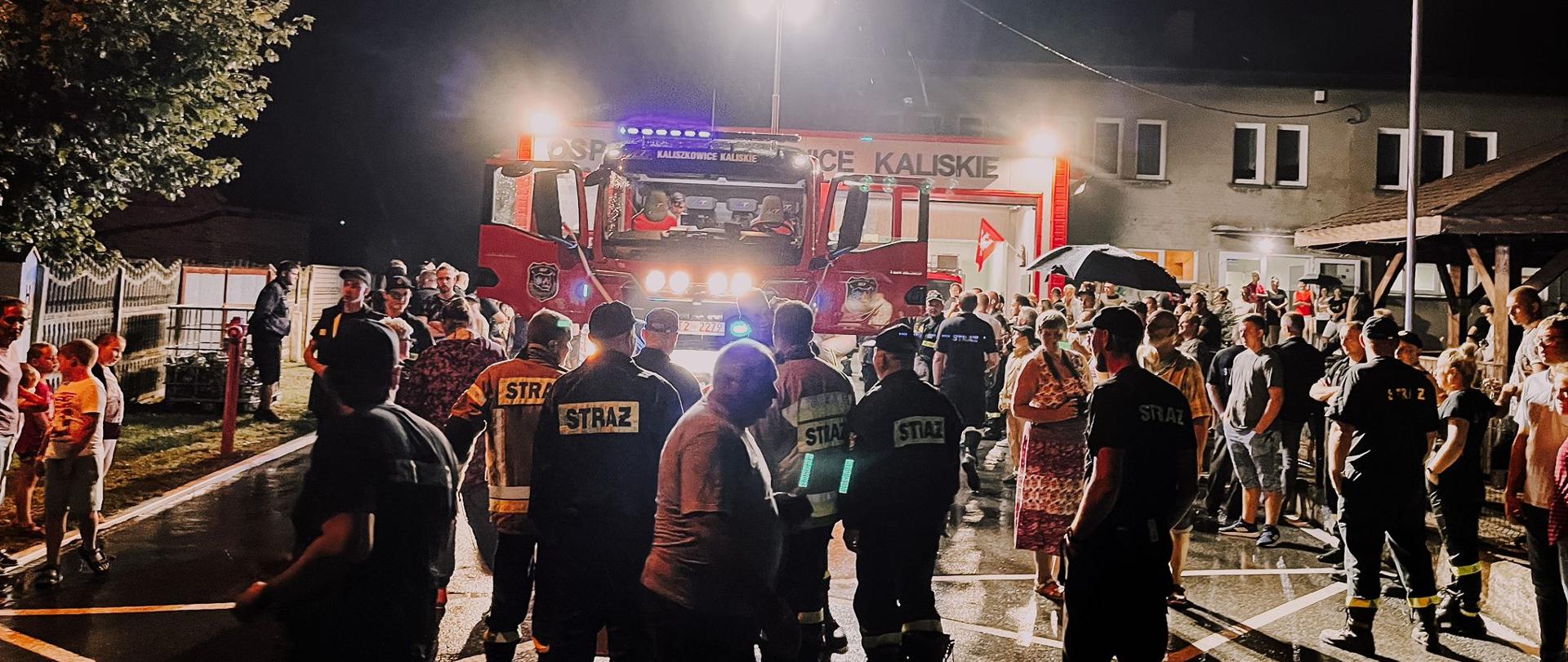 Na zdjęciu stoją osoby wokół nowego samochodu strażackiego, który stoi przed remizą. Zdjęcie wykonane porą nocną.