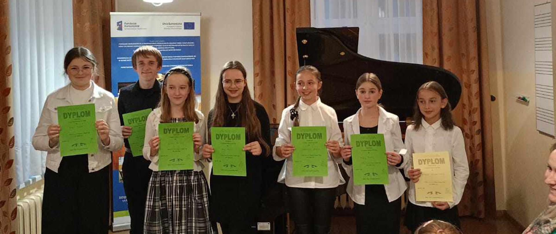 Zdjęcie w auli. Na pierwszym planie krzesła, kilka pustych na pozostałych siedzi publiczność. Następnie Uczniowie stoją w jednym rzędzie trzymający przed sobą dyplomy uczestnictwa i wyróżnienia. Za nimi fortepian, baner unijny oraz dwa okna a w nich firany oraz zasłony. Z lewej strony pod oknem grzejnik. Powyżej trzy zapalone, wiszące lampy