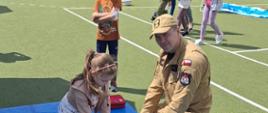 Na zdjęciu widać dorosłego mężczyznę w mundurze koloru khaki, strażaka, klęczącego na niebieskiej macie na sztucznej, zielonej trawie. Mężczyzna uczy małą dziewczynkę, również klęczącą, jak wykonywać uciśnięcia klatki piersiowej na manekinie do resuscytacji. Obok nich, nieco z tyłu, stoi chłopiec w pomarańczowej koszulce, obserwując. W tle widać wiele innych osób, głównie dzieci i dorosłych, na boisku sportowym, sugerującym wydarzenie plenerowe, takie jak piknik edukacyjny czy festyn. Z tyłu widać również siatki, namioty i inne elementy wyposażenia sportowego. W prawym dolnym rogu zdjęcia leży butelka wody.