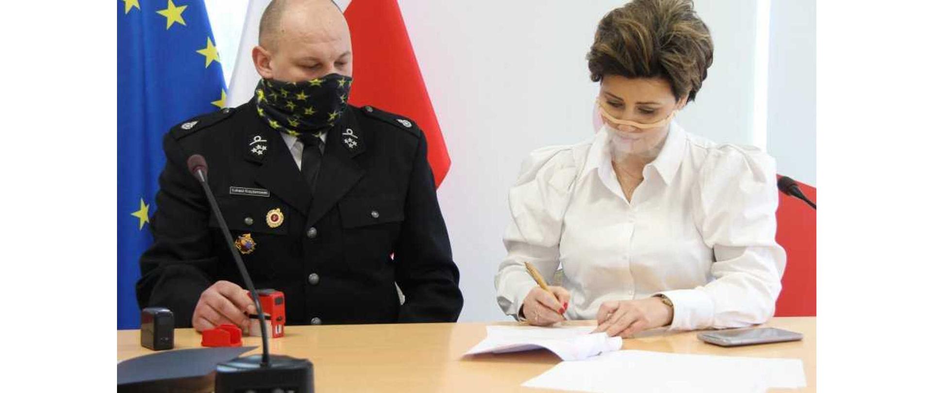 Strażak z OSP Skarżyn oraz Burmistrz Białej Piskiej siedzący przy biurku. Burmistrz Białej Piskiej podpisuje dokumenty dotyczące przekazania sprzętu ratowniczego jednostce OSP.