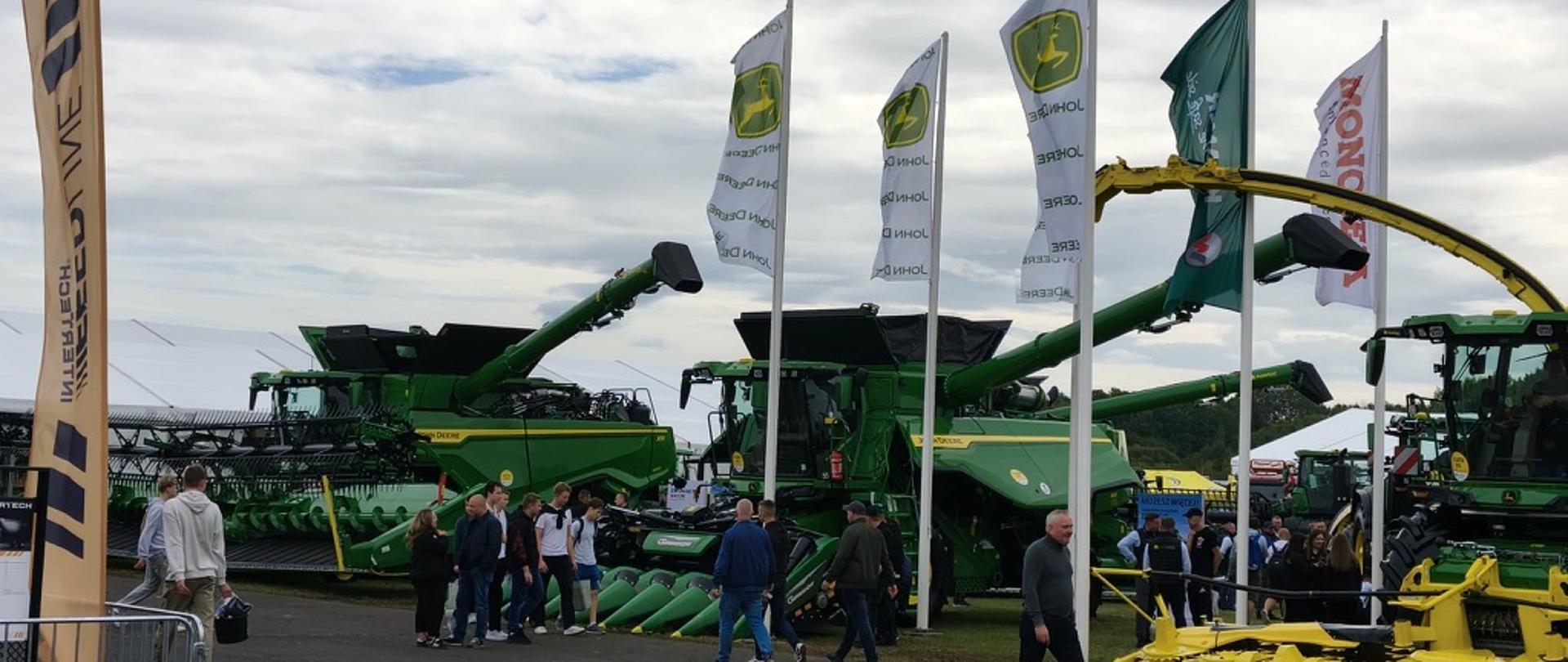 Kilka zielonych kombajnów zbożowych John Deere z rozłożonymi rurami wyładowczymi, na pierwszym planie flagi wystawcy. Po prawej sieczkarnia z żółtym zespołem żniwnym, przed maszynami spacerują zwiedzający.