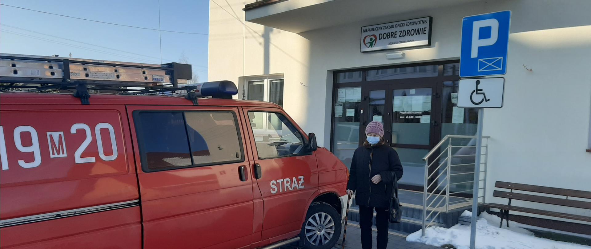 Starsza kobieta stojąca przy samochodzie OSP Rudzienko zaparkowanym przed przychodnią lekarską.