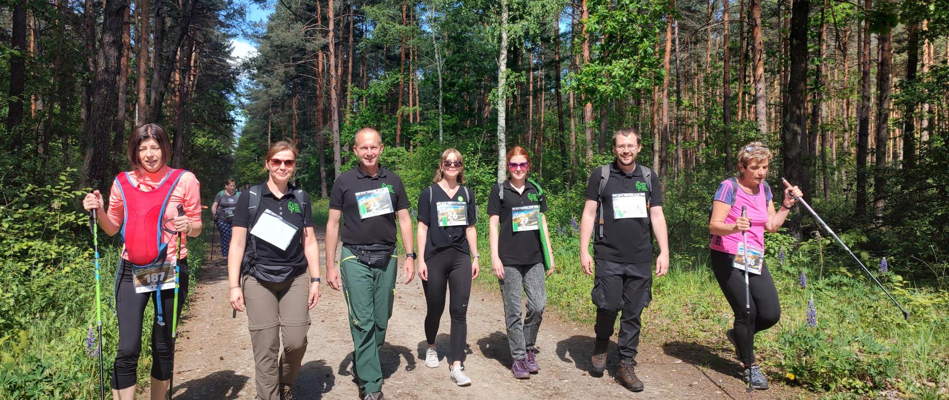 pięć osób w czarnych koszulach i dwie kobiety z kijkami nordic walking w trakcie spaceru przez las