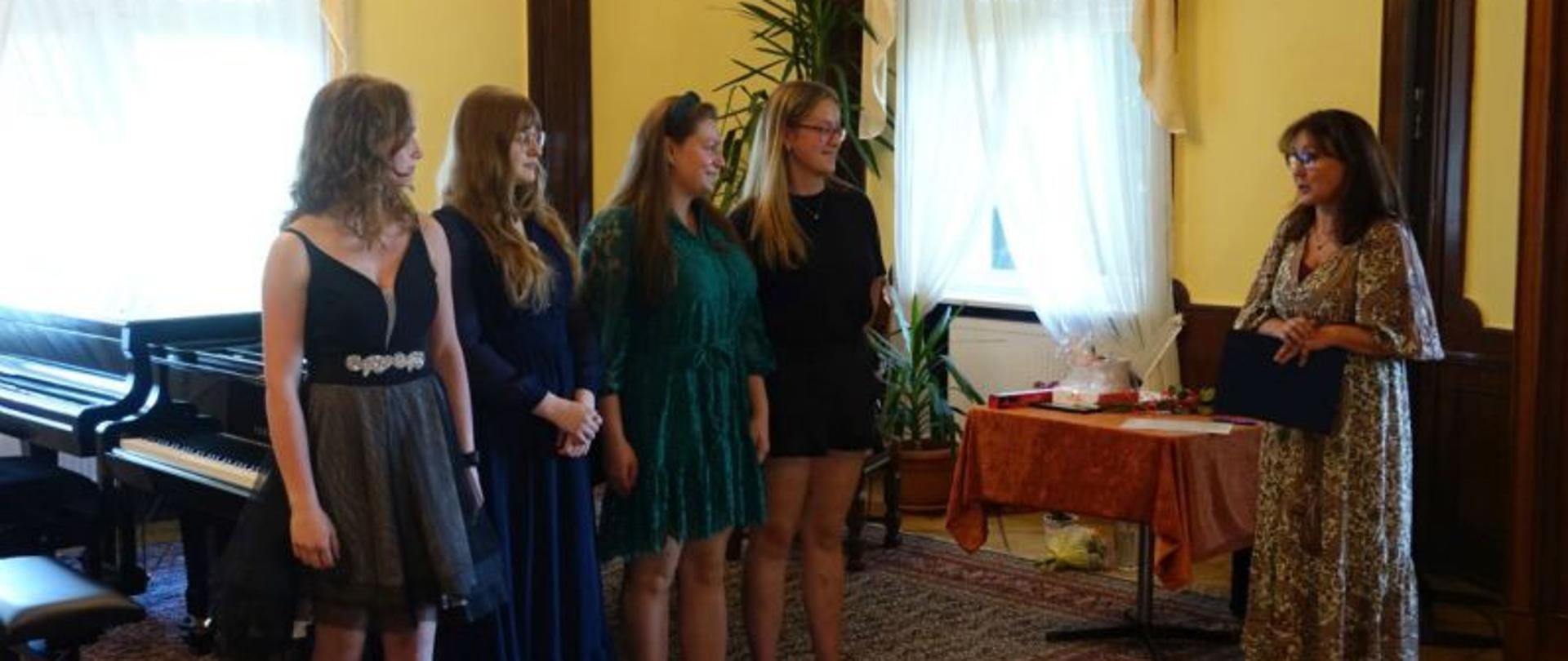 Zdjęcie przedstawiające absolwentów PSM w Słupsku stojących na tle fortepianu w sali kameralnej PSM w Słupsku. Na zdjęciu od lewej Amelia Smolińska, Ada Treder, Klaudia Kozakiewicz, Alicja Michalak. Po prawej Dyrektor PSM w Słupsku - Beata Podgórna.
