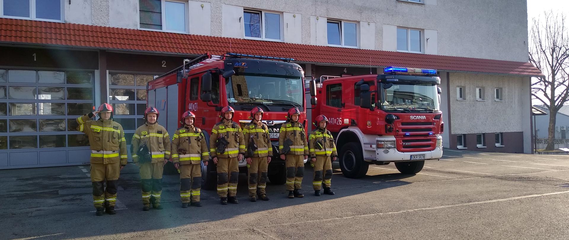 Na tle budynku JRG ustawione dwa pojazdy pożarnicze z włączonymi światłami alarmowymi. Przed samochodami w szeregu stoi siedmiu strażaków w ubraniach specjalnych i hełmach, Stojący po lewej stronie dowódca zmiany salutuje. Zdjęcie zrobione ze skosu od lewej strony szeregu. 