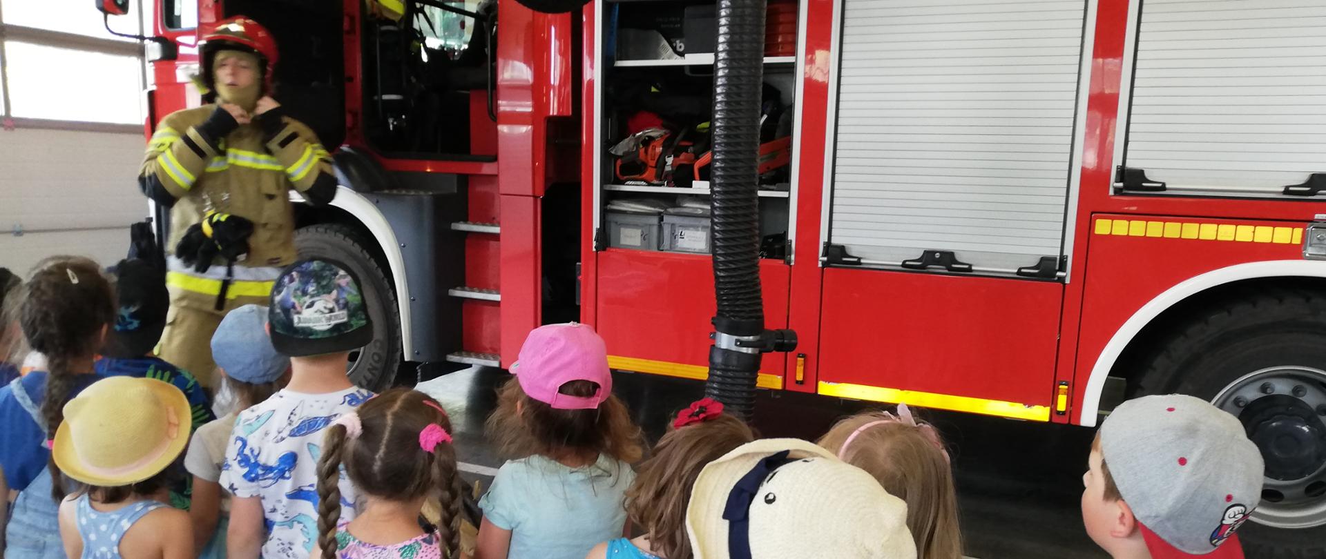 grupka dzieci obserwuje pokaz ubrania specjalnego, w tle samochód pożarniczy