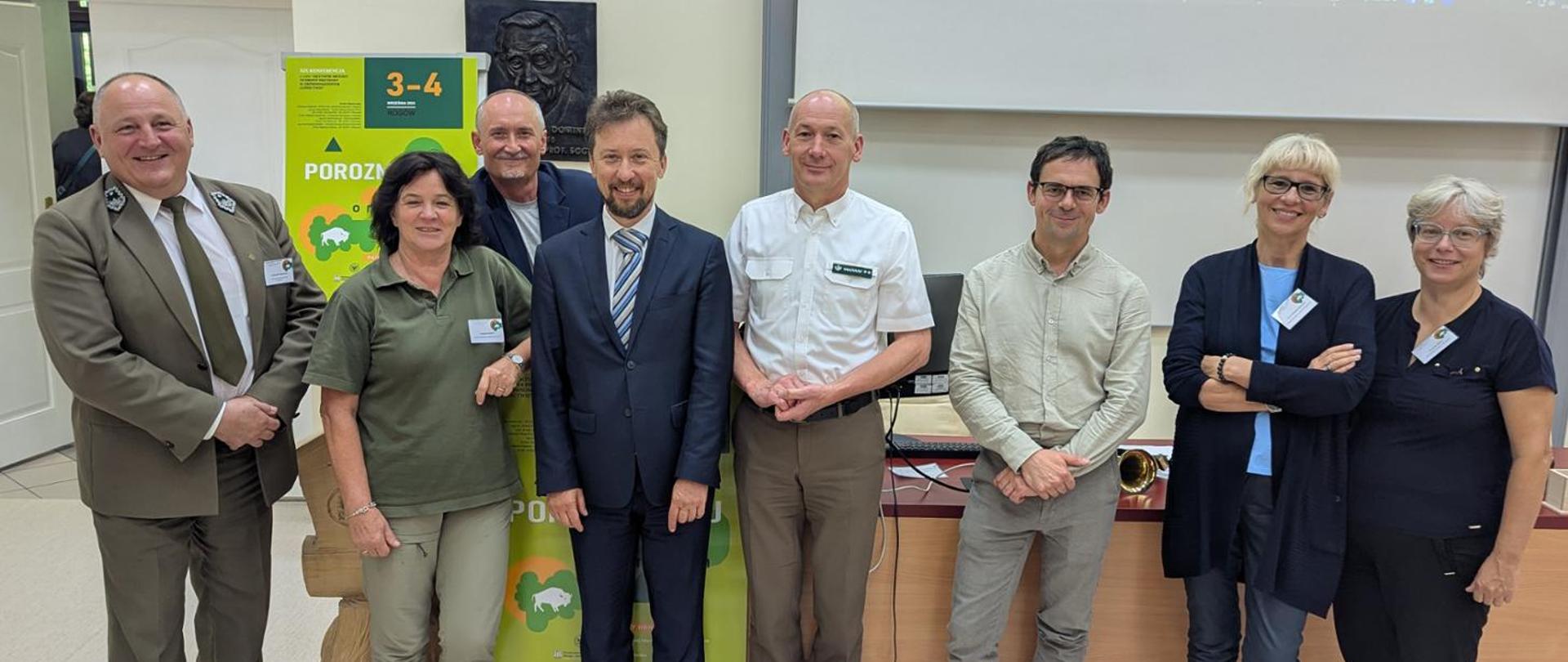 8 osób patrzy z usmiechem w obiektyw. Za nimi cześciowo widoczy roll-up i biała tablica