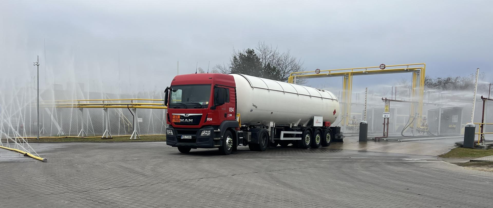 Na pierwszym planie stoi biała cysterna, a po prawej instalacje chłodzone mgłą wodną. Z przodu zdjęcia widoczny jest zielony trawnik, a powyżej pochmurne niebo. 