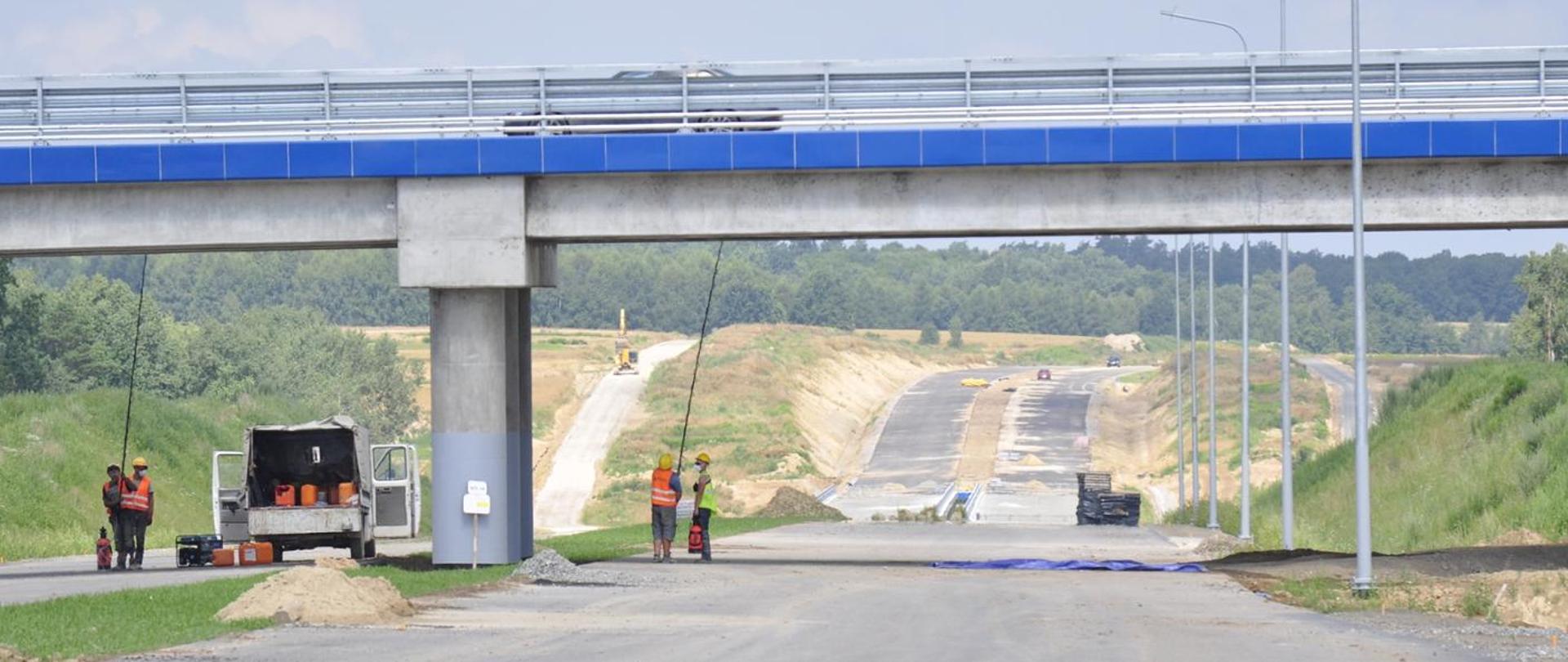 Robotnicy drogowi pracują pod betonowym wiaduktem na budowie nowej drogi. Na pierwszym planie stoi otwarta skrzynia samochodu serwisowego, a w tle widać nieukończony odcinek trasy prowadzącej przez wykop. Na placu budowy znajdują się maszyny i materiały, a całość otaczają zielone skarpy i drzewa.