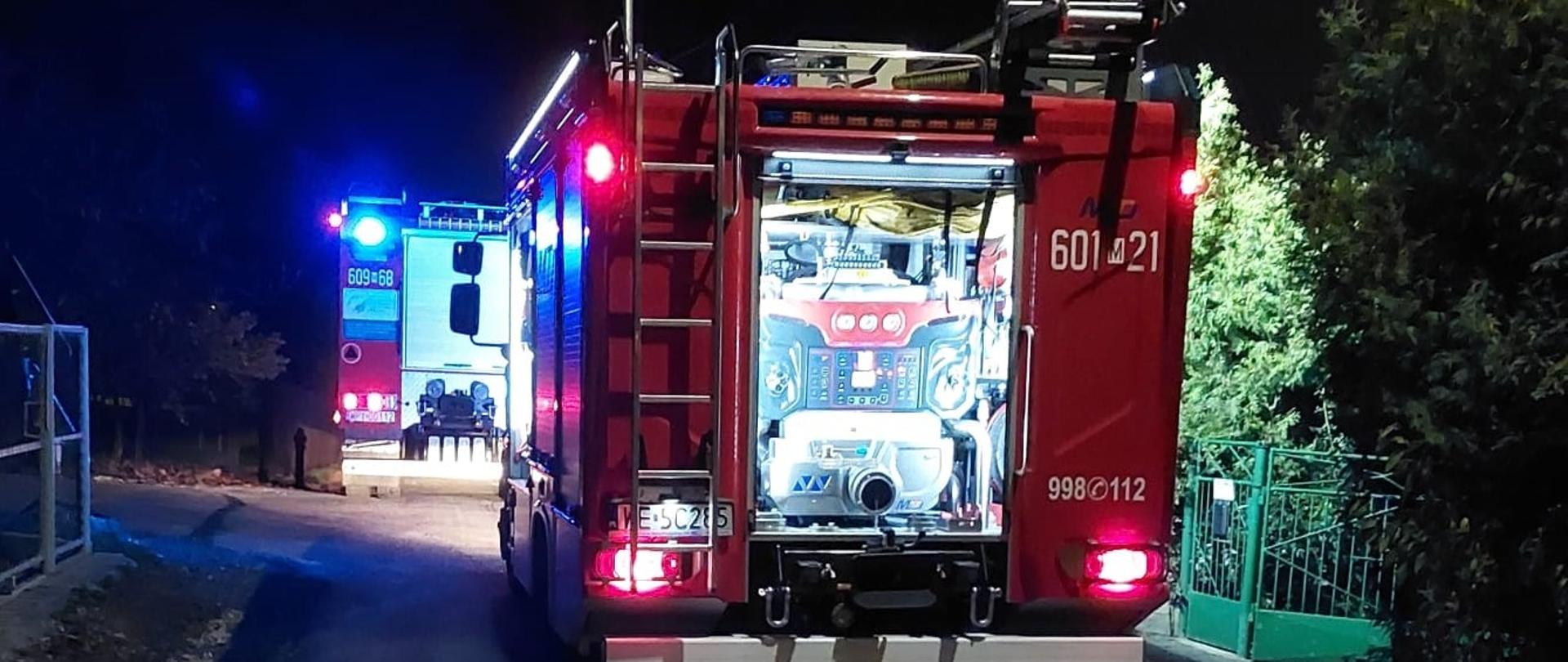 Zdjęcie przedstawia tył czerwonego wozu strażackiego z numerem "601 21" oraz numerami alarmowymi "998 112", zaparkowanego na wąskiej drodze, prawdopodobnie dojazdowej do posesji. Scena rozgrywa się w nocy, co podkreśla ciemne otoczenie i włączone światła wozów. Na dachu wozu widać wysunięty maszt oświetleniowy, emitujący jasne światło. Z tyłu pojazdu widoczne są drabiny oraz sprzęt gaśniczy. W oddali, za pierwszym wozem, widać kolejny pojazd uprzywilejowany, prawdopodobnie karetkę lub inny wóz strażacki, również z włączonymi niebieskimi światłami sygnalizacyjnymi. Na drodze, tuż za pierwszym wozem strażackim, widoczna jest plama na asfalcie, odbijająca czerwone światło od pojazdu. Po prawej stronie drogi widoczny jest fragment zielonego ogrodzenia i ciemne drzewa, natomiast po lewej stronie znajduje się siatkowe ogrodzenie i krzewy. Całość sugeruje, że na miejscu odbywa się nocna interwencja służb ratunkowych.