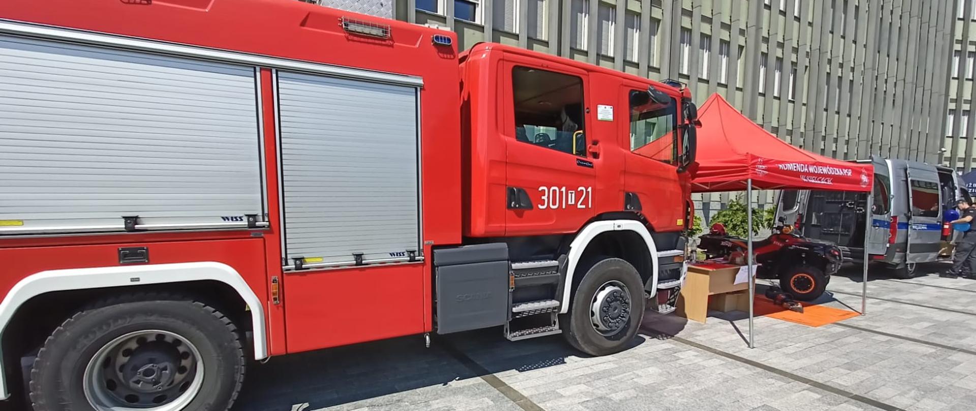 Zdjęcie przedstawia stoisko Państwowej Straży Pożarnej pod budynkiem świętokrzyskiego Urzędu Wojewódzkiego: od lewej samochód gaśniczy, namiot z stoiskiem i zaparkowany strażacki quad.