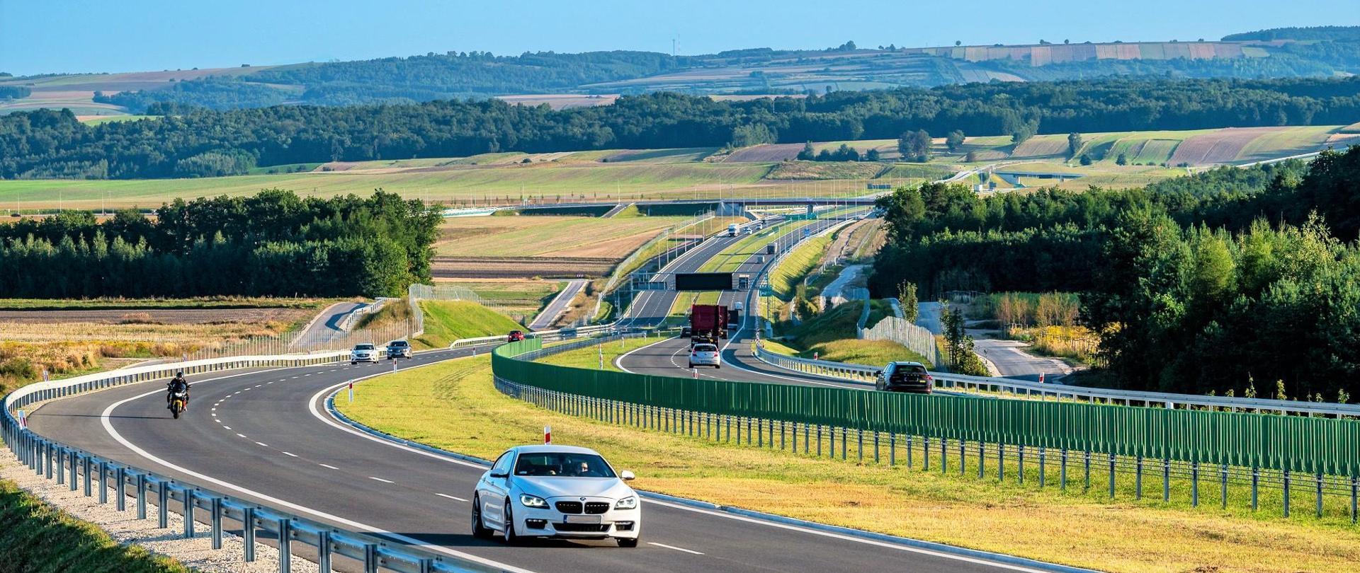 Droga ekspresowa S7 na odcinku Moczydło - Miechów. Dwujezdniowa droga ekspresowa wijąca się między polami i lasami po niewielkich pagórkach. Widoczne pojazdy poruszające się w obu kierunkach.