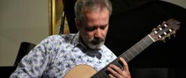 W pomieszczeniu. Pan Kamil Bartnik siedzi i gra na gitarze klasycznej. W tle czarne fortepian.