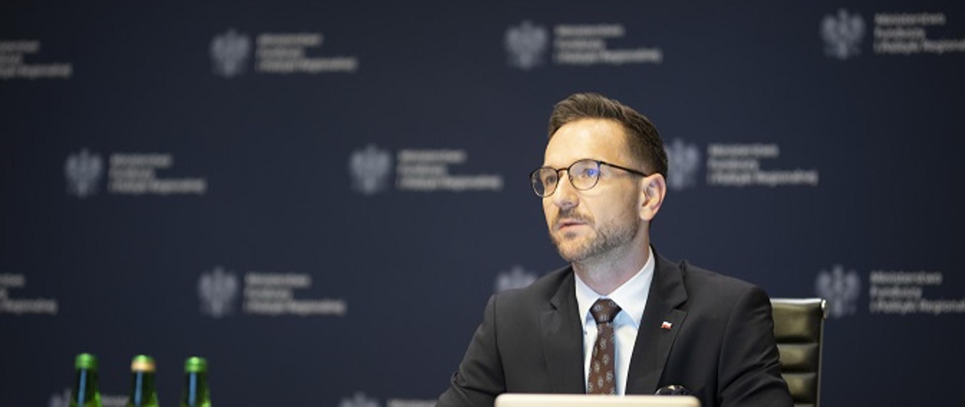 Wiceminister Waldemar Buda siedzi za stołem konferencyjnym, za nim tło z logo MFiPR