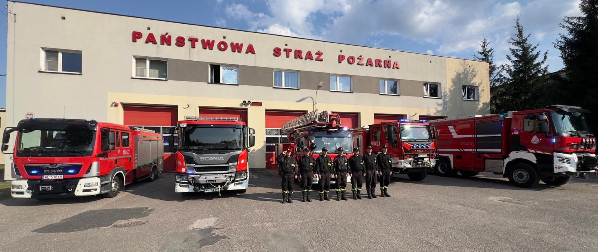 strażacy stojący na placu przed budynkiem komendy, za nimi stoją samochody pożarnicze