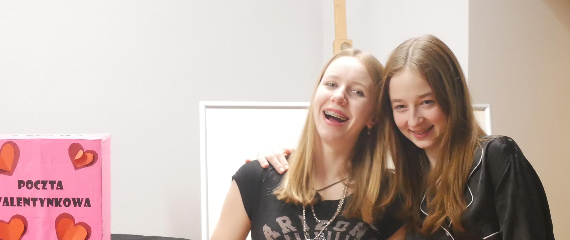 Dwie uśmiechnięte wychowanki stoją obok fortepianu z miskami pełnymi cukierków. Obok nich na fortepianie jest różowe pudełko z czerwonymi serduszkami. W pudełku są kartki walentynkowe.