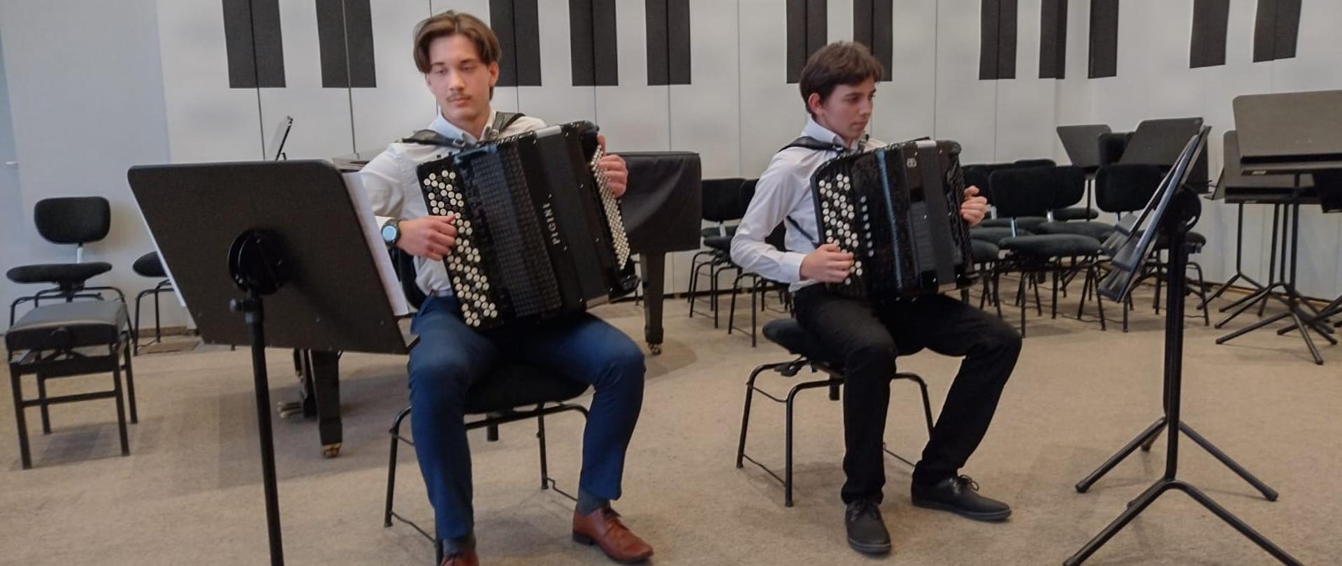 Dwaj starsi chłopcy siedzą na krzesłach i grają na akordeonach, po bokach z lewej u prawej strony stoją pulpity.