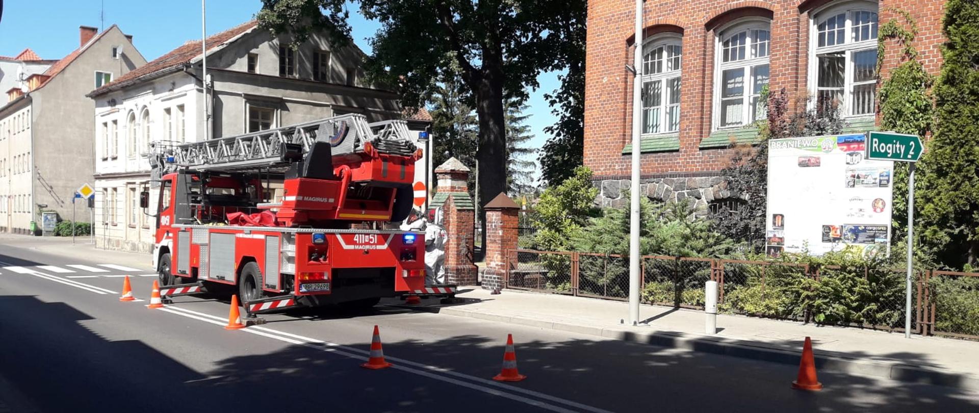 Samochód drabina strażacka ustawiana na drodze, ogrodzona pomarańczowymi pachołkami. W tle budynki.