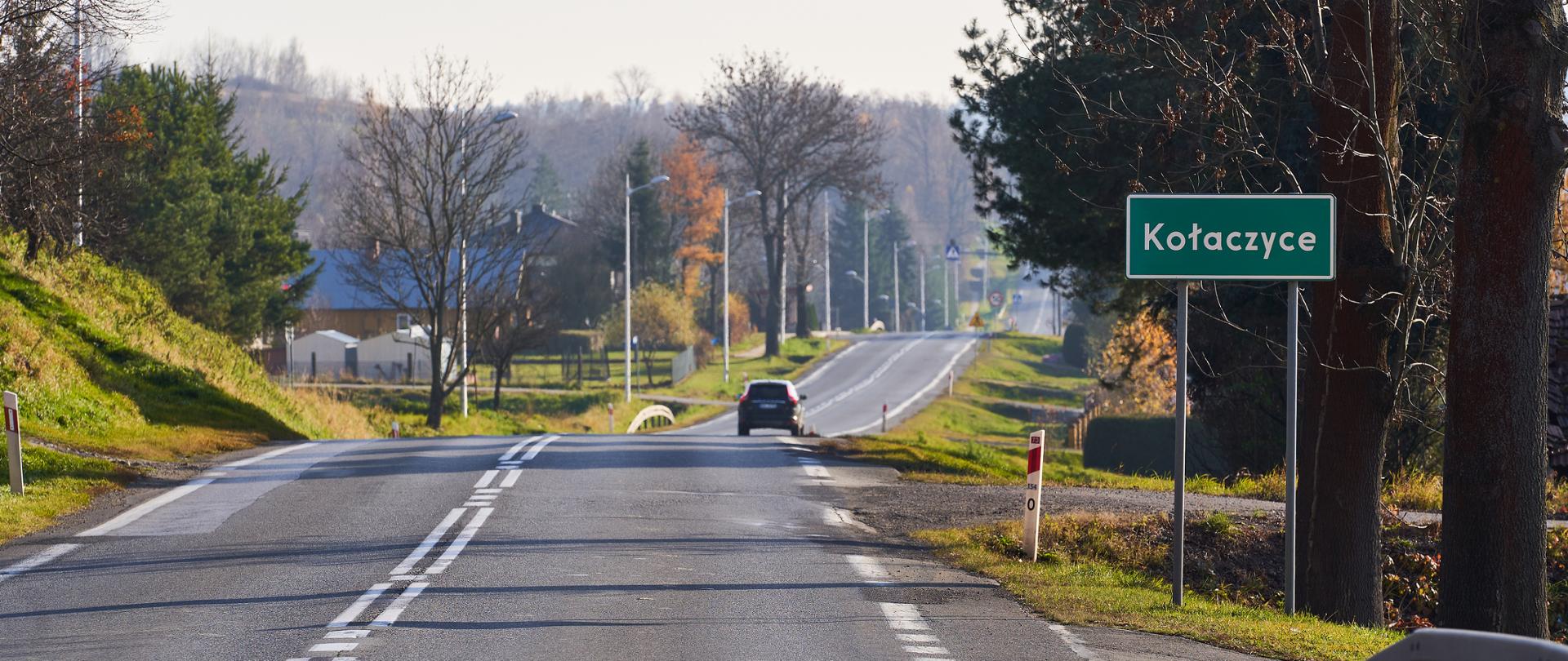 Widok na dwupasmową drogę biegnącą przez niewielką miejscowość. Po prawej stronie stoi zielona tablica z nazwą „Kołaczyce”. Wzdłuż drogi rosną drzewa, widać pojedyncze domy, latarnie uliczne oraz samochód jadący w oddali. Słoneczny dzień późną jesienią.