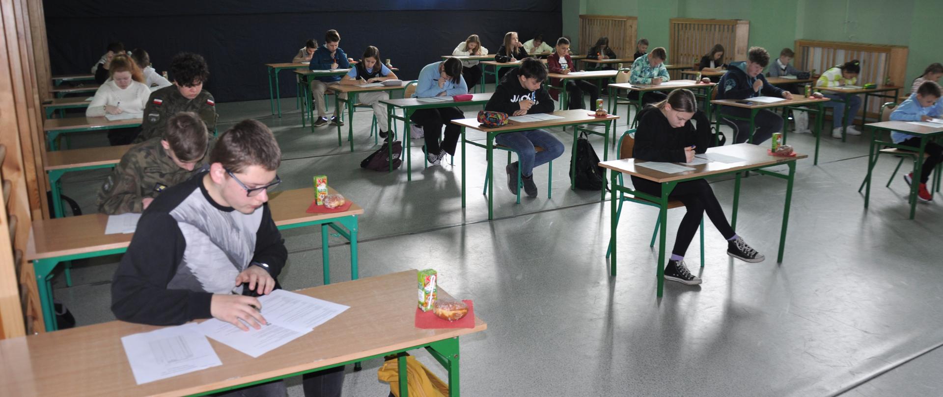 Sala szkoły podstawowej z ławkami a za nimi uczniowie piszący test OTWP