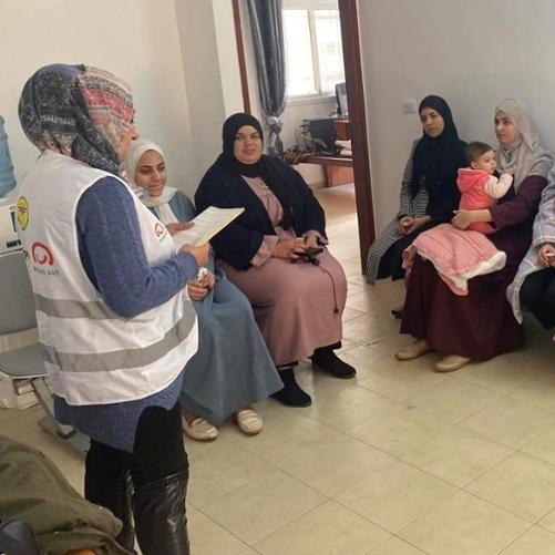 Kobiety z głowami ubranymi w chusty siedzą na krzesłach pod ścianą. Przed nimi stoi kobieta, również z głową zasłoniętą chustą i patrzy się na kartki papieru, które trzyma przed sobą. 