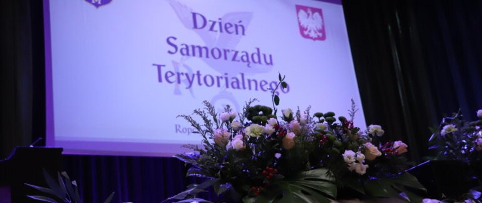 Zdjęcie przedstawiające slajd z napisem Dzień Samorządu terytorialnego oraz wiązankę kwiatów. 