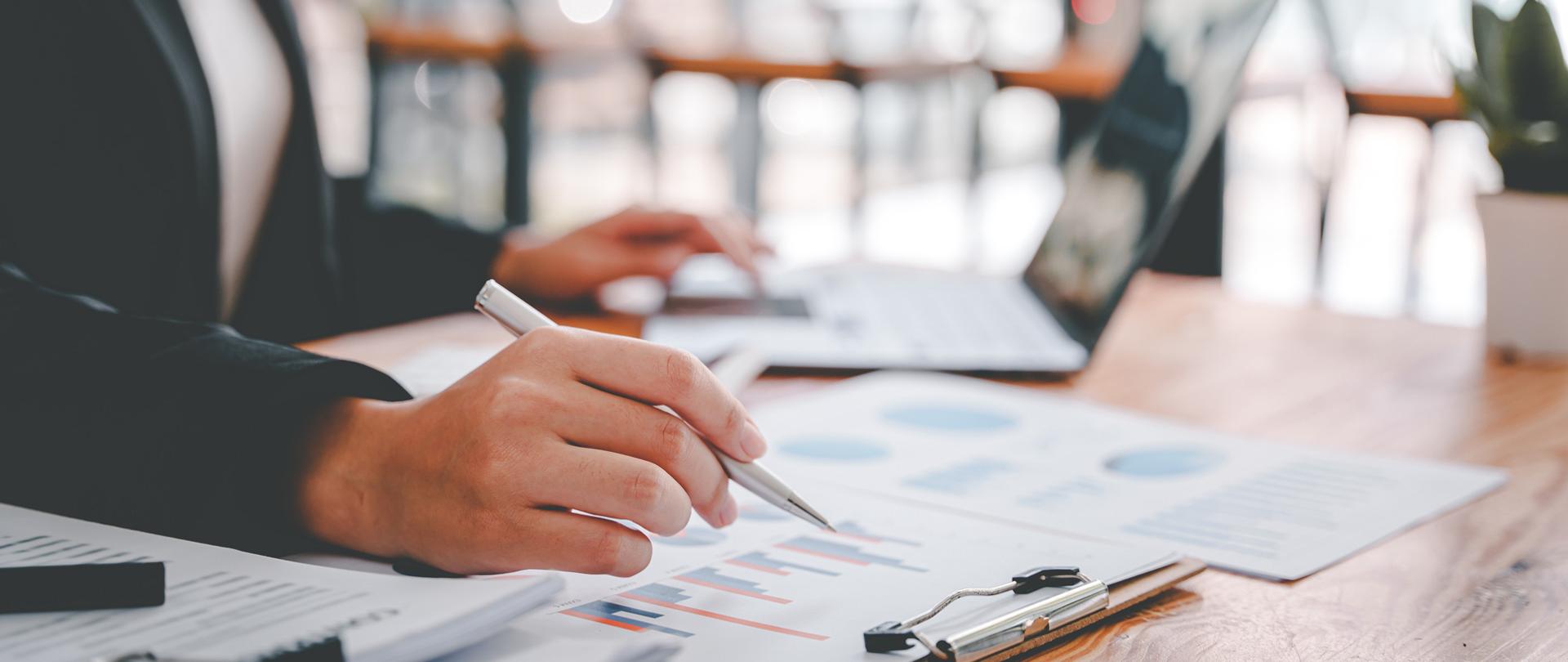 Dłoń trzymająca długopis, dokumenty i komputer na biurku.