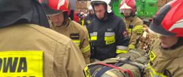 Strażacy niosą kukłę pozoranta podczas ćwiczeń. W tle skład drewna oraz maszyny z tartaku.